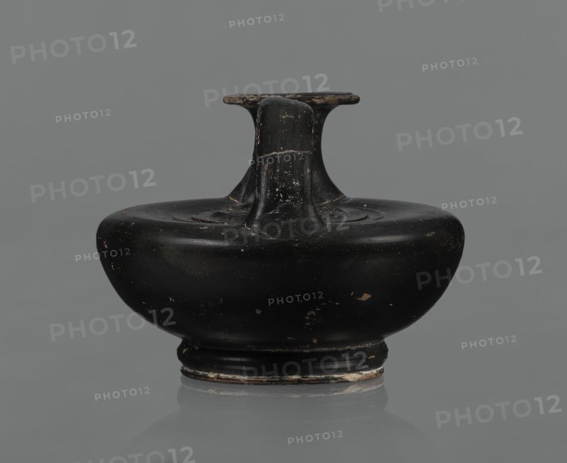 Black-finished askos, unknown Campanian workshop, black-finished pottery, askos, Campania Italy. Askos with a profiled foot and a semicircular body closed at the top with a slightly recessed surface. Narrow neck ending in a wide rim. Ribbon-shaped handle attached to the neck and to the upper surface of the vessel. Appliqué with a female head on the upper surface, probably Medusa, surrounded by a wreath of intertwined snakes. Finish is black, shiny, worn in places, particularly on the handle and the rim edge. Light orange clay. Handle and neck cracked and glued. Mid 3rd century BC., A black rounded ceramic vessel with a short neck and a flat, T-shaped rim sits centered against a neutral gray background; the surface shows worn areas with small chips and lighter scratches revealing a tan or brownish underlayer, and the base rim has patches of lighter wear.