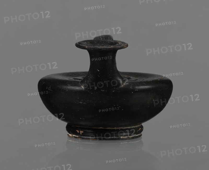 Black-finished askos, unknown Campanian workshop, black-finished pottery, askos, Campania Italy. Askos with a profiled foot and a semicircular body closed at the top with a slightly recessed surface. Narrow neck ending in a wide rim. Ribbon-shaped handle attached to the neck and to the upper surface of the vessel. Appliqué of a female head glued to the upper surface of the vessel, probably Medusa, surrounded by a wreath of intertwined snakes. Finish is black, shiny, worn in places, particularly on the handle and the edge of the rim. Light orange clay. Handle and neck cracked and glued. Mid 3rd century BC., A small rounded ceramic vessel with a wide flattened body and a short flared neck topped by a flat disc-like rim, shown against a plain neutral background; the surface is predominantly black with visible small areas of worn lighter color and slight scuffs, and the base shows a few patches of lighter wear; the overall shape is symmetrical and compact with smooth curved contours and a ...