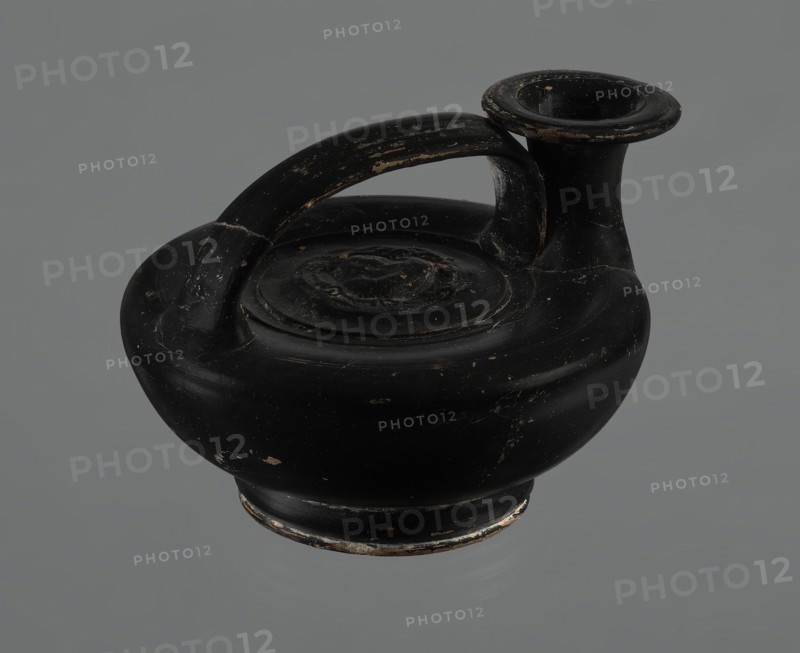 Black-finished askos, unknown Campanian workshop, black-finished pottery, askos, Campania Italy, Askos with a profiled foot and a semicircular body closed at the top with a slightly recessed surface. Narrow neck ending in a wide rim. Ribbon-shaped handle attached to the neck and to the upper surface of the vessel. Appliqué with a female head glued to the upper surface of the vessel. The head depicted is probably Medusa, surrounded by a wreath of intertwined snakes. The finish is black, shiny, worn in places, particularly on the handle and the edge of the rim. Light orange clay. Handle and neck cracked and glued. Mid 3rd century BCE, Campania, vessel., A black rounded ceramic vessel with a single curved handle arching over the top and a short flared spout, showing worn areas with small patches of lighter color and surface scratches, sitting against a plain gray background.