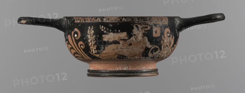 Red-figure bowl with Eros and a young woman; red-figure pottery, bowl kylix; 3rd quarter of the 4th century BC; Apulia. Yellow clay tending to red; glossy black engobe; some elements painted white. Bowl with a ring-shaped base with two vices attached to the rim horizontally; palmettes beneath the vices. Belly decorated with two scenes: Scene A — young woman semi-reclining on the ground, facing left, holding a wreath in her left hand and a woven basket on her outstretched right hand; dressed in a long, smooth chiton; neck adorned with a pearl necklace; wrists with double bracelets; hair tied in a bun with a kekryphalos. Scene B — Eros flying, facing left; in the right hand a musical instrument and in the left hand a wreath with a ribbon; hairstyle and jewelry like Scene A bun with kekryphalos, double bracelets, pearl necklace; ankles with bracelets and head with a diadem; entire figure has hermaphrodite features. Condition: numerous chips; engobe dull in places. Dimensions: height 5.1 c...