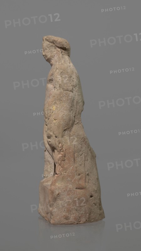 Terracotta figurine of Heracles, terracotta, figurines of nude men, Heracles mythology, Greek mythology, figurines made of molds. Figurine of a standing naked Heracles in a contrapposto pose with a cloak thrown over his back. His right hand rests on a rock, and in his left he probably holds a club. Condition of preservation: good. 2nd–1st century BC, Asia Minor, figurine, height 14.3 cm, A worn stone figurine of a standing person shown in profile on a neutral gray background, the figure carved with a rounded head, a suggestion of facial features, and a short hairstyle or head covering; the body appears clothed with a draped form extending to a square base, showing surface weathering, small pits, and abrasions; the stone displays tones of beige, tan, light brown, pale pink, and traces of yellow and greenish discoloration, with faint white veining and patches of darker brown.
