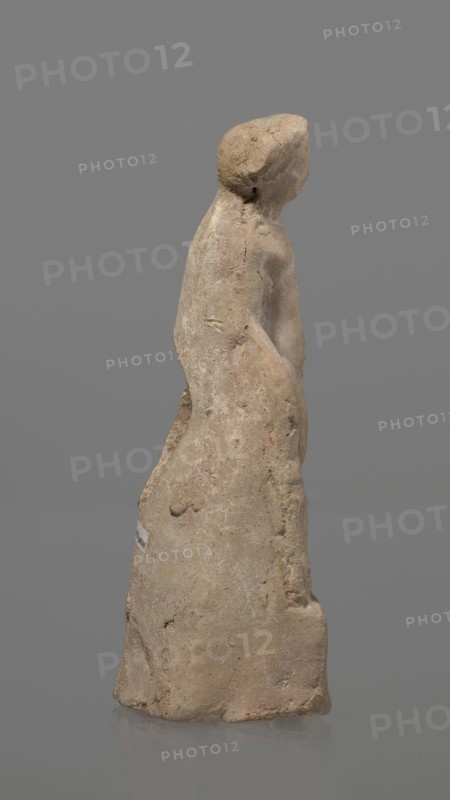 Terracotta figurine of Heracles. Figurine of a standing naked Heracles in a contrapposto pose with a cloak thrown over his back. His right hand rests on a rock, and in his left he probably holds a club. Terracotta, made from a mold. Figurine of a nude male, Greek mythology. Condition: good. Date: 2nd–1st century BC. Place of origin: Asia Minor. Height: 14.3 cm., A roughly carved standing figure of a human form made of a light tan, beige material with areas of darker brown and faint yellowish spots, showing a simplified head, shoulders, and torso; the surface is textured with visible pitting and small cracks, and there are worn, smoother areas on the torso and head. The figure is viewed in profile, with one arm suggested against the body and the lower portion flaring into a skirt-like shape; the base is uneven and rests on a smooth reflective surface. The background is a uniform medium gray.