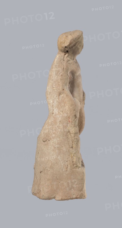 Terracotta figurine of Heracles, terracotta, figurines of nude men, Heracles mythology, Greek mythology, figurines made of molds. Figurine of a standing naked Heracles in a contrapposto pose. He has a cloak thrown over his back. His right hand rests on a rock, and in his left he probably holds a club. Condition of preservation: good. 2nd–1st century BC, Asia Minor, figurine, height 14.3 cm., A weathered clay figure viewed in profile against a plain gray background, with an elongated, rounded head and a simplified facial plane, a slender neck leading to sloping shoulders and an elongated torso that tapers into a long skirt-like lower form; the surface is rough and pitted with irregular texture and small chips, and the figure has a prominent rounded protrusion at the chest and an arm that hangs close to the body with an indistinct hand; visible colors include muted beige, light tan, pale brown, and hints of ochre and gray on the surface.