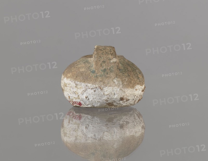 Oil lamp, Islamic art, Persian, utility ceramic for everyday use. Oil lamp with a round container with a flattened bottom, a strongly elongated and flatly cut burner, and a rounded handle. The upper part of the lamp is covered with green glaze. 12th century, Iran. Height 4.4 cm, width 9.8 cm, depth 6.0 cm., A rounded, weathered object with an irregular short protrusion on top sits centered against a plain gray background, its surface showing a rough texture with areas of brown, beige, white, and patches of green, and a faint red mark near the lower edge; the object is reflected on a smooth reflective surface below, mirroring the same colors and textures.