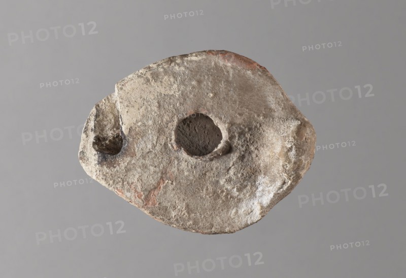 Oil lamp, maker, Islamic art; Arabic culture; oil lamp; Arabic pottery; utility pottery; everyday life in the Middle East; conical handle; traceably marked foot; 8th–10th century, Middle East; height 5.8 cm, width 11.8 cm, depth 9.1 cm, A roughly oval, weathered object with a coarse, pitted surface sits against a plain background; its colors include pale beige, light gray, off-white, and patches of reddish-brown, with darker brown visible inside two irregular depressions, one central and one toward an edge; the surface shows rough texture, small pits, and subtle raised areas, and the overall shape is asymmetrical with uneven edges and worn contours.