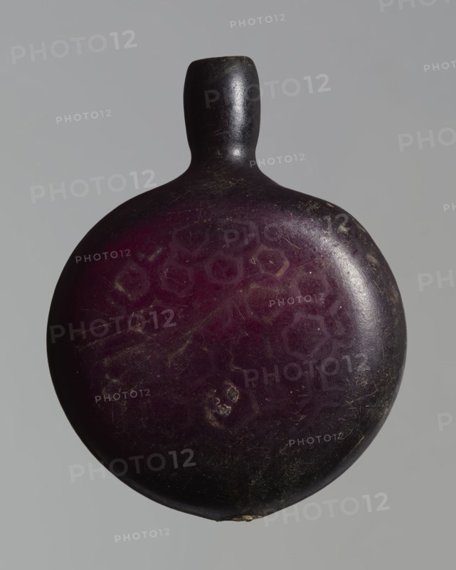 Pilgrim's bottle, Islamic/Arabic, Ayyubid dynasty 12th–13th century, Syria/Egypt/Middle East probable origin. Violet manganese glass. The belly is widened at the arms, which merge into a cylindrical, slightly convex neck with a narrow opening. One wall bears a star motif with a rosette in the middle and a palmette; the other and the side arms show a honeycomb motif. Vessel; height 11.8 cm, width 9.1 cm, depth 4.2 cm., A rounded object with a short neck and rounded top, shown against a plain gray background, with an overall deep purple surface that transitions to dark brown and black toward the edges; the surface shows a few lighter purple hexagonal patterns, several small scratches and scuffs, and a few pale worn spots.
