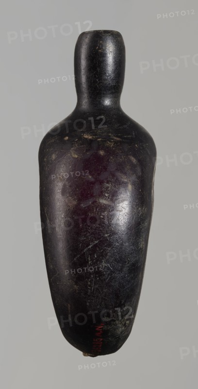 Pilgrim's bottle, gift; Islamic art, Arabic/Islamic culture; Ayyubid dynasty 12th–13th century. The belly is widened at the arms, which merge into a cylindrical, slightly convex neck with a narrow opening. One wall has a star motif with a rosette in the middle and a palmette; the other wall and the side arms have a honeycomb motif. Violet manganese glass. Syria/Egypt/Middle East probable origin. Vessel. Height 11.8 cm; width 9.1 cm; depth 4.2 cm., A smooth, elongated object with a rounded top and tapered bottom resembling a bottle or pestle, shown against a plain light gray background; the object's surface is dark, appearing black with areas of deep purple and muted brownish patches, and it has a worn, slightly glossy texture with visible scratches and scuffs.