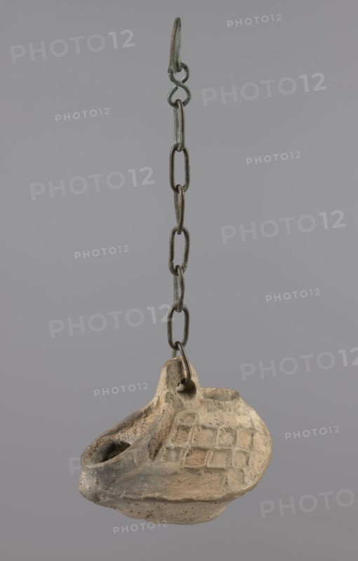 Lamp, Oval lamp made of yellow-gray clay with a high container and two handles, through which a bronze chain is strung for hanging the lamp. The high, sloping arms are decorated with a plastic ornament of a rectangular mesh. A channel running perpendicularly through the arms from the handle reaches the wick hole and surrounds it, creating a high rim. Coptic oil lamp, Egypt, 5th–6th century. Height 10 cm, width 7.1 cm., A hanging object suspended from a metal chain with a hook is visible against a plain gray background; the chain is dark metal with greenish patina and links of varying shapes, and attached at the bottom is a small worn vessel made of light beige clay with darker brown and gray discoloration, showing a rounded body, a spout-like opening, a raised area with a grid-like pattern of square impressions, and a small loop where it connects to the chain.