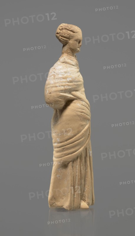 Terracotta figurine of a standing woman. Terracotta, molded figurine, Tanagra figurine. Figurine of a standing woman dressed in a chiton and himation. The body pose is in contrapposto. The hands are hidden under the robe — the left lowered, the right bent at the elbow and resting on the back. Hair tied in a bun. Missing polychrome and base. 4th century BC. Tanagra Greece. Figurine. Height 16.5 cm, width 6.5 cm, depth 4 cm., A standing sculpted figure viewed in profile, made of a light tan material with areas of pale discoloration, showing a wrapped garment with draped folds and a rounded shoulder, the head turned slightly forward with stylized hair gathered at the back, set against a solid medium gray background.