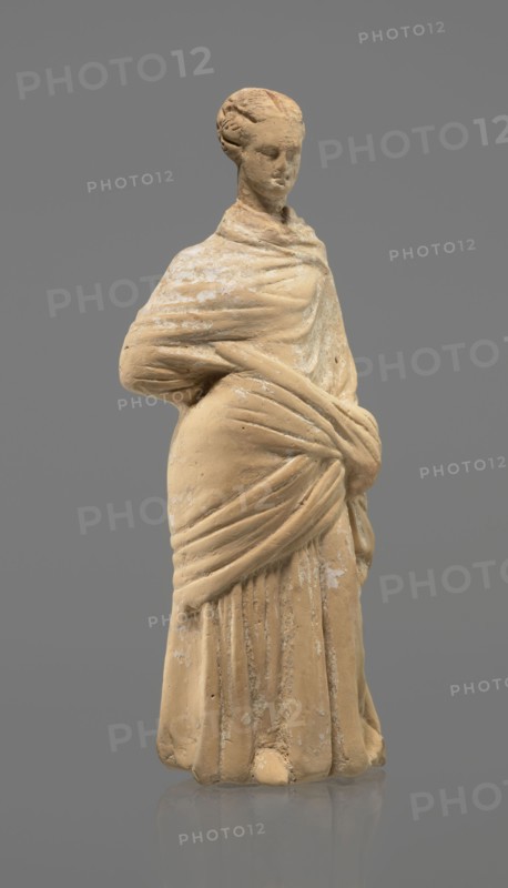 Terracotta figurine of a standing woman, terracotta, female figurine, himation, chiton, Tanagra figurine, molded, figurine of a standing woman dressed in a chiton and himation. The body pose is in contrapposto. The hands are hidden under the robe — the left lowered, the right bent at the elbow and resting on the back. Hair tied in a bun. Preservation condition: missing polychrome and base. 4th century BC, Tanagra Greece, figurine; height 16.5 cm, width 6.5 cm, depth 4 cm., A standing statue of a person draped in layered cloth, depicted with a wrapped garment that covers the shoulders and is gathered at the waist, showing sculpted folds and drapery. The figure has a braided or coiled hairstyle and a serene facial expression with closed or downcast eyes. The surface shows worn areas and patches of lighter residue against the main material. The colors visible are beige or tan for the statue material, lighter off-white patches on the surface, and a gray background.