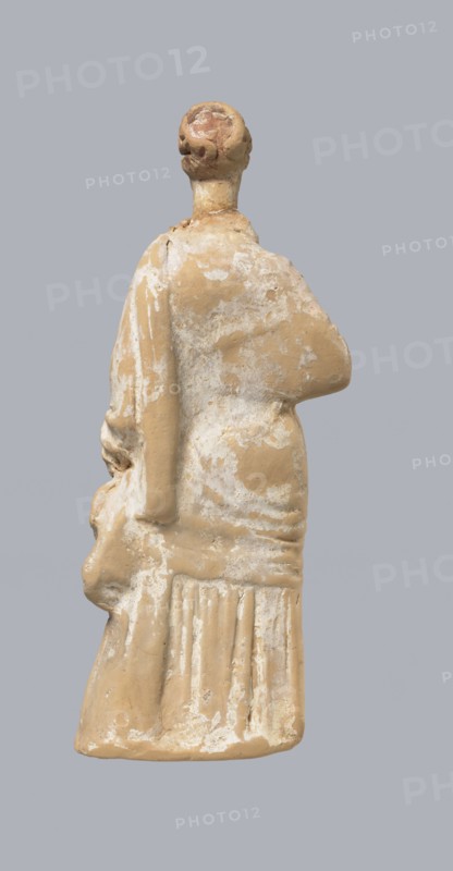 Terracotta figurine of a standing woman, terracotta, female figurine, himation, chiton, Tanagra figurine, molded figurine. Figurine of a standing woman dressed in a chiton and himation. The body pose is in contrapposto. The hands are hidden under the robe - the left one is lowered, the right one is bent at the elbow and rests on the back. Hair is tied in a bun. Preservation condition: missing polychrome and base. 4th century BC, Tanagra Greece, figurine, height 16.5 cm, width 6.5 cm, depth 4 cm, A weathered beige figurine viewed from behind against a light gray background, showing a rounded head with textured hair, a long neck, a draped upper garment with worn surface revealing lighter patches, one arm bent across the torso and the other hanging down holding a rounded object, and a lower skirt with vertical folds and scuffed lighter areas; visible colors include beige, light tan, off-white, and light gray.