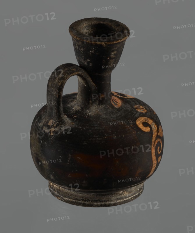 Red-figure aryballos lekythos, unknown Campanian workshop. A squat lekythos with a ring-shaped foot. The belly is broad, spherical, and flattened; the shoulders are rounded and broad. The narrow, low neck is as high as the rim. The rim is cup-shaped. The handle is ribbon-shaped, vertical. The front is decorated with a bar ornament at the base of the neck. The belly and arms depict a seated woman facing left: legs in profile left extended, right withdrawn, hips three-quarters en face, arms frontal, head in left profile. The left arm is wrapped around a himation, the hand resting on the hip; the right hand is outstretched, above which lies a tossed circular object of two light-colored parts ball or skein of wool?, each with a single black dot and separated by a dark stripe. Hair combed back and tied in a bun; woman wears a necklace and spherical-bead earrings depicted with dots. Sides feature vertically unwinding plant tendrils with spirally coiled ends. Reddish clay; turning lines visib...