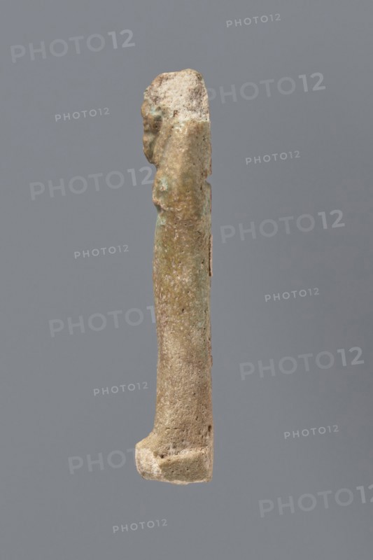 Ushabti, mummy-shaped funerary figurine ushabti; gift inscription by Meyet, L.; materials not specified; Late Period, 525–343 BC 27th–30th Dynasty, Egypt; height 8.7 cm, A slender vertical object photographed against a solid gray background, showing a weathered surface with a rough, pitted texture and irregular contours; the object is primarily beige and light brown with areas of pale green and pale bluish-green patina, and small darker brown spots; the top appears more irregular and slightly chunkier while the lower end broadens into a thicker base with similar rough texture; lighting is soft, casting minimal shadow and revealing subtle surface variations.