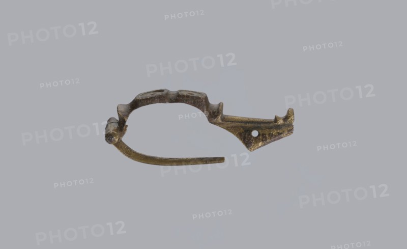 Arched fibula, unknown Roman workshop. Fibula with an arched hoop decorated with two crossbars and rectangular plates with embossed circles. The pin grows from a cylindrical crossbar. The long, flat sheath is decorated with an oval plate on the front. Preservation condition: parts of the sheath and pin are missing. Date: 75–150, Roman Empire 27 BC–476. Material: bronze. Type: fibula. Dimensions: height 1.6 cm, width 4.1 cm, weight 0.006 kg., An irregular metallic object with a curved, open loop shape rests against a plain light gray background; the object appears tarnished with a mix of dull gold, brown, and dark gray tones, showing rough and pitted surfaces, a hinge-like thicker section at one end, a tapered pointed extension with a small round perforation toward the other end, and a thin curved arm projecting from the loop.