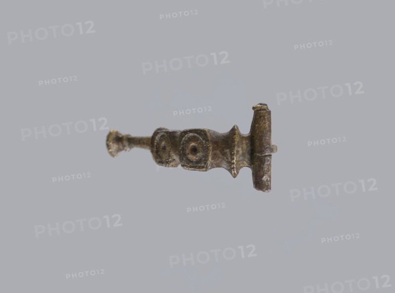 Arched fibula, unknown Roman workshop. Fibula with an arched hoop decorated with two crossbars and rectangular plates with embossed circles. The pin grows from a cylindrical crossbar. The long, flat sheath is decorated with an oval plate on the front. Preservation condition: parts of the sheath and pin are missing. Date: 75–150, Roman Empire 27 BC–476. Material: bronze. Type: fibula. Dimensions: height 1.6 cm, width 4.1 cm, weight 0.006 kg., A small metallic object suspended against a pale gray background, showing a horizontal rod-like shape with textured surfaces and irregular contours; the object appears bronze or brass-toned with areas of darker brown and hints of greenish patina, featuring a thicker end with a rounded vertical post and a narrower flared end, and a central section with two adjacent raised circular motifs and ribbed ridges along its profile.