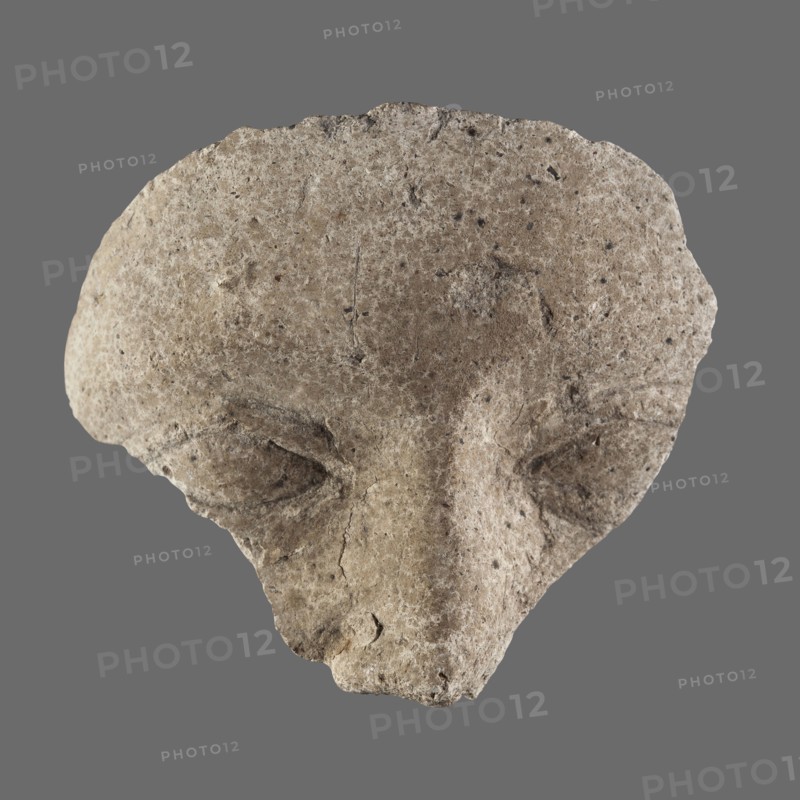 Fragment of a stucco mask; upper part of a sarcophagus mask showing forehead, eyes and nose. Sarcophagus mask, stucco, height 8.8 cm, width 8.9 cm. 2nd century, Roman period, Egypt., A roughly triangular, weathered stone fragment with a textured, pitted surface appears against a flat gray background; the stone shows a palette of beige, tan, light brown, and specks of darker brown and black, with areas of lighter off-white highlights; the shape suggests shallow carved depressions resembling eye sockets and a raised central ridge resembling a nose, with irregular edges, small cracks, and rough granular detail across the surface.