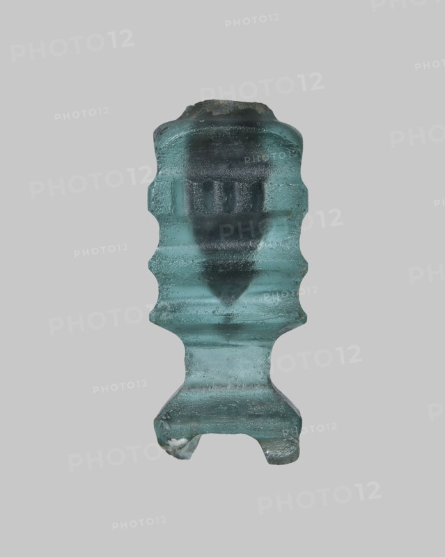 Cosmetic bottle, ; Islamic, Arabic culture; 9th–11th century, Egypt. Thick-walled blue glass vessel, height 4.1 cm, width 1.7 cm. On the body one convex band decorated with fine horizontal grooves; flat arms; heavily carved base with four feet; wheel-cut decoration., A single translucent object with a textured surface centered against a plain light gray background, showing shades of aqua, teal, and darker green-blue; the object has an irregular vertical shape with a rounded top, constricted midsections creating wavy contours, and a broader lower portion that forms a short pedestal with two small protrusions at the base; surface details include visible ridges, smooth and rough patches, and variations in translucency producing darker internal areas and lighter outer highlights.