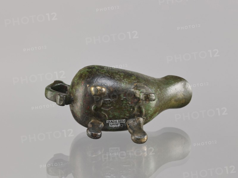 Yi-type vessel, Chinese culture, Chinese art, ceremonial ritual vessel, Qing dynasty 1644–1911, 19th century, China, vessel on four legs one broken off; underneath, an image of a human, cult object, height 6.4 cm, width 11.0 cm, depth 4.5 cm., A small rounded sculptural object rests on a reflective surface, showing a patinated finish with visible green and dark brown areas and spots of brighter bronze where the surface is worn; it has a bulbous body tapering into a narrower rounded end, with a looped handle on one side and several short stumpy protrusions on the underside; the backdrop is a smooth neutral gray, and the object casts a soft reflected silhouette on the surface beneath it.