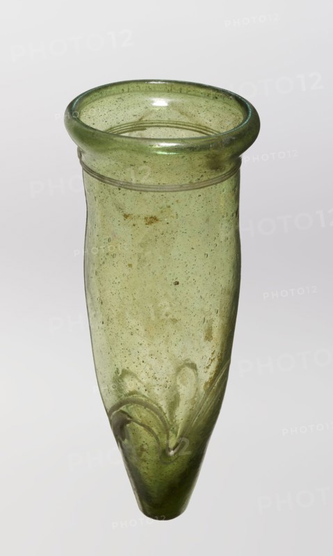 Lamp oil container, glass with cream enamel thread, 12th–15th century, possibly Syria or Egypt Middle East, Islamic/Arabic, oil lamp, height 12.0 cm, diameter 5.2 cm., A translucent green glass vessel with a rounded flared rim and a long tapering body, showing a bubbly texture and some cloudy inclusions throughout; the rim is a darker green band while the body is a lighter yellow-green with subtle brownish spots and streaks, and near the pointed end there are raised swirl-like ridges in the glass; the background is plain and pale.