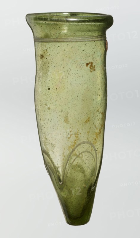 Lamp oil container, deposit provenance, Islamic art, Arabic culture, glass oil lamp, cream enamel thread also at the bottom, 12th–15th century, Syria, or Egypt, Middle East, height 12.0 cm, diameter 5.2 cm, A tall, narrow glass vessel with a rounded flared rim and a tapered pointed base, rendered in translucent green with areas of pale and deeper green tones; the surface shows many small bubbles and a few irregular darker spots and patches, plus a few rusty brown discolorations; there is a subtle applied horizontal ridge near the rim and a decorative wavy applied thread near the lower body creating arched shapes; overall the glass has a slightly uneven, hand-blown texture and a glossy finish.