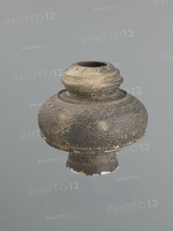 Jug, Persian, Islamic art; 16th–18th century, Central Asia ; ceramic vessel with bulging belly on a high foot; decoration of plant twig and raised geometric and geometric-floral patterns; height 18.7 cm, width 18.3 cm;, A rounded ceramic vessel with a short neck and flared base sits against a plain pale gray background; the vessel is predominantly dark gray to black with areas of lighter gray and beige weathering, and it has carved or incised swirling decorative patterns and bands of raised ridges around the neck and shoulder, with small spots of white chips and worn edges along the base.
