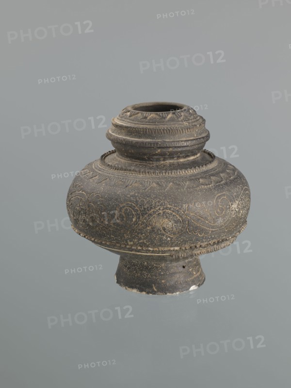 Jug, Persian, 16th–18th century, Central Asia, earthenware vessel with bulging belly on a high foot, height 18.7 cm, width 18.3 cm. Decoration: plant twig and raised geometric and geometric-floral ornament., An ornate round vessel with a flared base and a short neck, shown against a plain gray background; the object appears dark brown to black with lighter worn areas and fine incised decorative patterns of swirling lines and leaf-like motifs around its bulbous body, and textured horizontal bands and ridges around the neck and shoulder.