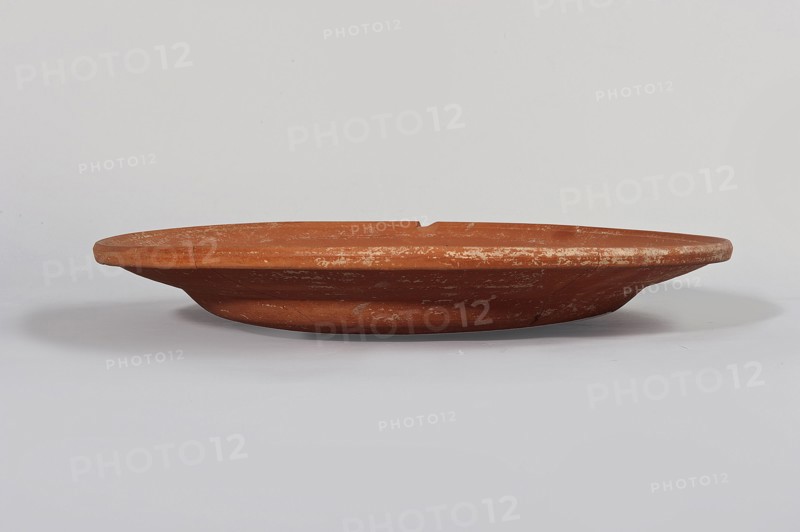 Terra sigillata plate. Flat plate with a very low foot and a rather flat, wide rim. Made of clay with orange glaze. 2nd half of the 4th century. Found in Tunisia. Plate, diameter 33.5 cm, height 5.6 cm., A low, shallow terracotta dish with a wide, flat rim and a slightly raised base sits centered against a pale background; the dish shows a weathered surface with irregular pale patches and scratches, and its colors include reddish-brown, lighter beige scuffs, and subtle gray shadows on the underside and background.
