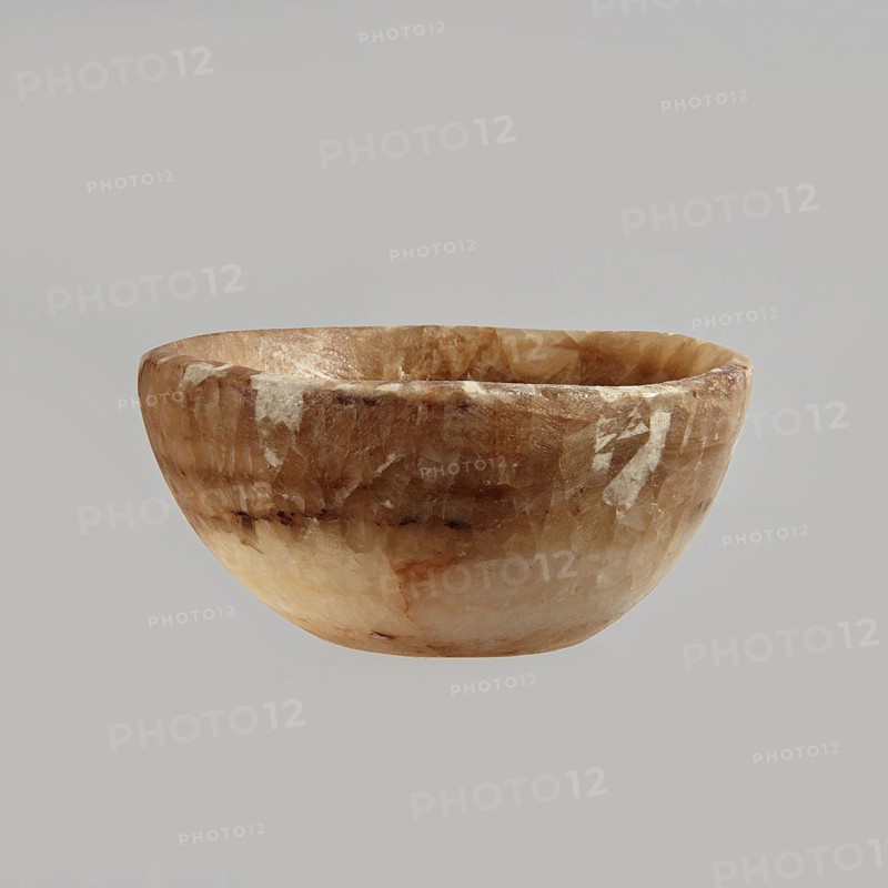 Bowl made of striped, coarse-crystalline Egyptian alabaster, waxed, with a flat bottom and gently sloping walls. 20th century, Egypt. Forgery. Vessel. Height 4 cm, width 9 cm., A rounded bowl with a smooth, slightly irregular rim sits against a plain background; the bowl's surface shows a marbled pattern of warm beige, light brown, creamy white, and hints of darker brown, with visible translucent and opaque patches and subtle variations in texture and sheen.