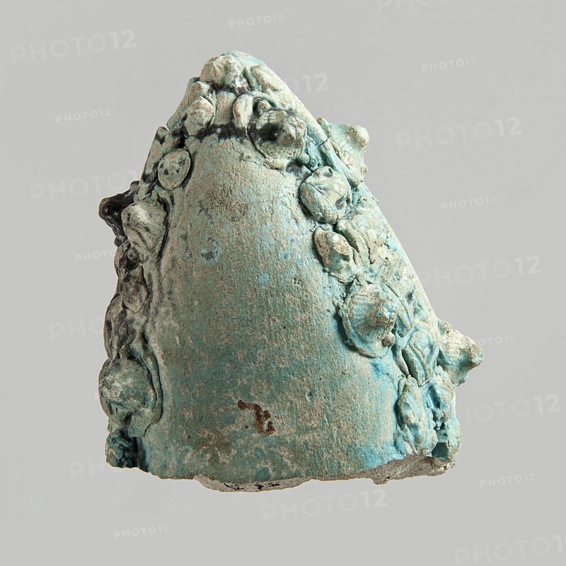 Fragment of a votive boat. Prow or stern with decoration in the form of glued appliqués. On the sides from the top: four lion heads. On the deck: palmette, fish, sitting cat with a disc above its back, cartouche half preserved. On the outside sides: two fish, four lion heads, four rosettes, three cats with a disc, two falcons with a uraeus behind their backs, four falcons with a neb symbol and a nechacha whip behind them one partially preserved, two en face faces, two rosettes one partially preserved. Bottom smooth, slightly convex. Blue glaze, discolored black in places. c. 30 BC–395 AD, Roman period, Egypt. Votive offering. Height 4.5 cm; width 11 cm; depth 8.9 cm., An irregular, cone-shaped object with a rough, textured surface covered in a patina of turquoise and pale green, with areas of darker blue-green and small patches of brown and beige; the object has a ring of rounded, lumpy protrusions clustered along its sides and top, and its base shows a thin, uneven edge with exposed g...