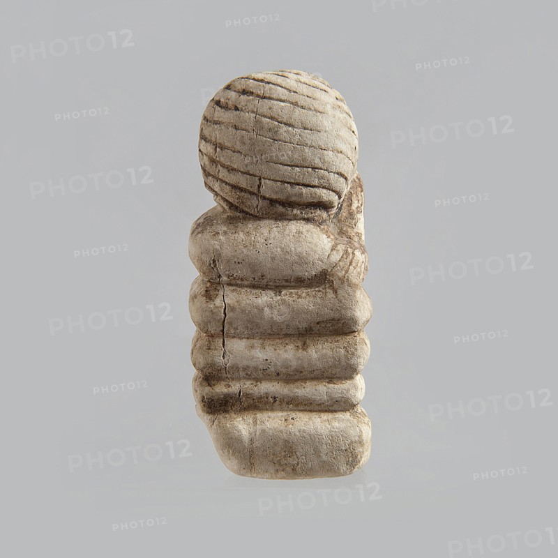 Fragment of a figurine. Head of a polychrome figurine, which originally probably depicted a woman lying on a bed with her head propped up on a pillow. The woman's head is shown wearing a short, striped wig. Behind the head, a woman's hand is visible, which, like the head, rests on an object that may represent a pillow or bolts of cloth. 332 BC–395 AD; Ptolemaic Period–Roman Period. Egypt. Figurine. Height 2.8 cm; width 5.6 cm; depth 2.5 cm., A small sculpted object sits against a plain light gray background, appearing in shades of beige, tan, and light brown with darker brown weathered lines and spots; the top portion is a rounded shape with carved horizontal grooves, and below it a stacked, segmented column of rounded rectangular forms shows visible cracks and textured surface wear, with shadows and subtle highlights accentuating the carved contours and irregularities.