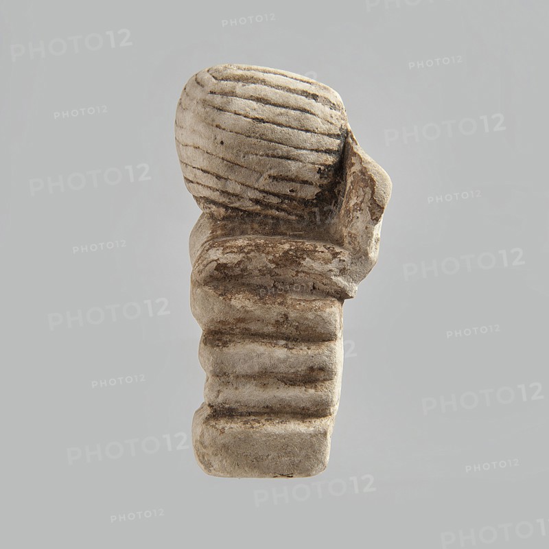 Head of a polychrome figurine, which originally probably depicted a woman lying on a bed with her head propped up on a pillow. The woman's head is shown wearing a short, striped wig. Behind the head, a woman's hand is visible, which, like the head, rests on an object that may represent a pillow or bolts of cloth. Fragment of a figurine. 332 BC–395 AD; Ptolemaic Period–Roman Period. Egypt. Figurine. Height 2.8 cm, width 5.6 cm, depth 2.5 cm., A weathered stone object with a rounded top featuring carved horizontal grooves, sitting on a stacked, ribbed base; the surface shows areas of rough texture and erosion with darker patches. The visible colors are shades of beige, light gray, and darker brown on a pale gray background.