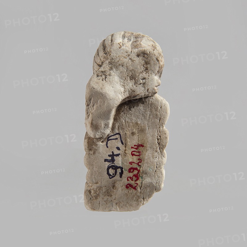 Head of a polychrome figurine, which originally probably depicted a woman lying on a bed with her head propped up on a pillow . The woman's head is shown wearing a short, striped wig. Behind the head, a woman's hand is visible, which, like the head, rests on an object that may represent a pillow or bolts of cloth. Fragment of a figurine. 332 BC–395 AD; Ptolemaic Period–Roman Period. Egypt. Figurine. Height 2.8 cm, width 5.6 cm, depth 2.5 cm., A weathered, pale stone fragment shaped like a small carved figure is centered against a plain light gray background, showing a rounded head with carved hair and a draped section flowing down the side; the stone surface displays a range of off-white, beige, tan, and light brown tones with darker brown weathering and small rough cracks and pits, and a few traces of darker markings on the stone's face and body.