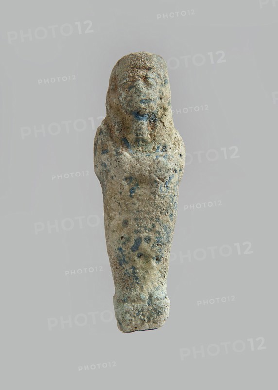 Ushabti. Carelessly made figurine with schematic facial features, in a narrow wig revealing the ears, with a rather short beard. In his hands he holds a mattock. The back is flat; the end of the wig is marked with an engraved line. Feet on a pedestal. Glaze almost completely worn away. Third Intermediate Period, Egypt. Ushabti; height 6.5 cm, width 2.1 cm, depth 1.3 cm., A small worn figurine with a tapered, elongated body and a rounded head, showing a rough, pitted surface texture with several irregular spots of darker coloration; the object displays muted beige and tan tones with areas of blue-green and gray mottling, and appears to have faint, shallow contours suggesting facial features and shoulder forms while the lower end is slightly flattened.