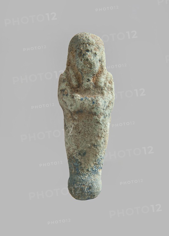 Ushabti. Carelessly made figurine with schematic facial features, in a narrow wig revealing the ears, with a rather short beard. In his hands he holds a mattock. The back is flat; the end of the wig is marked with an engraved line. Feet on a pedestal. Glaze almost completely worn away. Third Intermediate Period, Egypt. Ushabti; height 6.5 cm, width 2 cm, depth 1.4 cm. Maker and exact provenance unknown., A small weathered figurine shaped like a standing human figure with crossed arms, showing a rounded head and stylized facial features, with a narrow, tapered lower body and a rounded base; the surface appears rough and pitted with areas of erosion and encrustation, and the visible colors include pale beige, sandy brown, muted greenish-blue, and touches of darker bluish-gray.