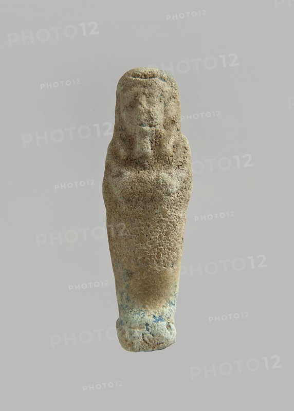Ushabti. Carelessly made figurine with schematic facial features, in a narrow wig revealing the ears, with a rather short beard. In his hands he holds a mattock. The back is flat, the end of the wig is marked with an engraved line. Feet on a pedestal. Glaze almost completely worn away. Third Intermediate Period, Egypt. Ushabti; height 6.3 cm, width 1.9 cm, depth 1.3 cm., A single upright carved figure with a textured, weathered surface in a range of beige and light brown tones with patches of pale green and faint blue, featuring a stylized head with a rounded top and minimal facial contours and a tapered body that narrows toward the base, set against a neutral gray background.