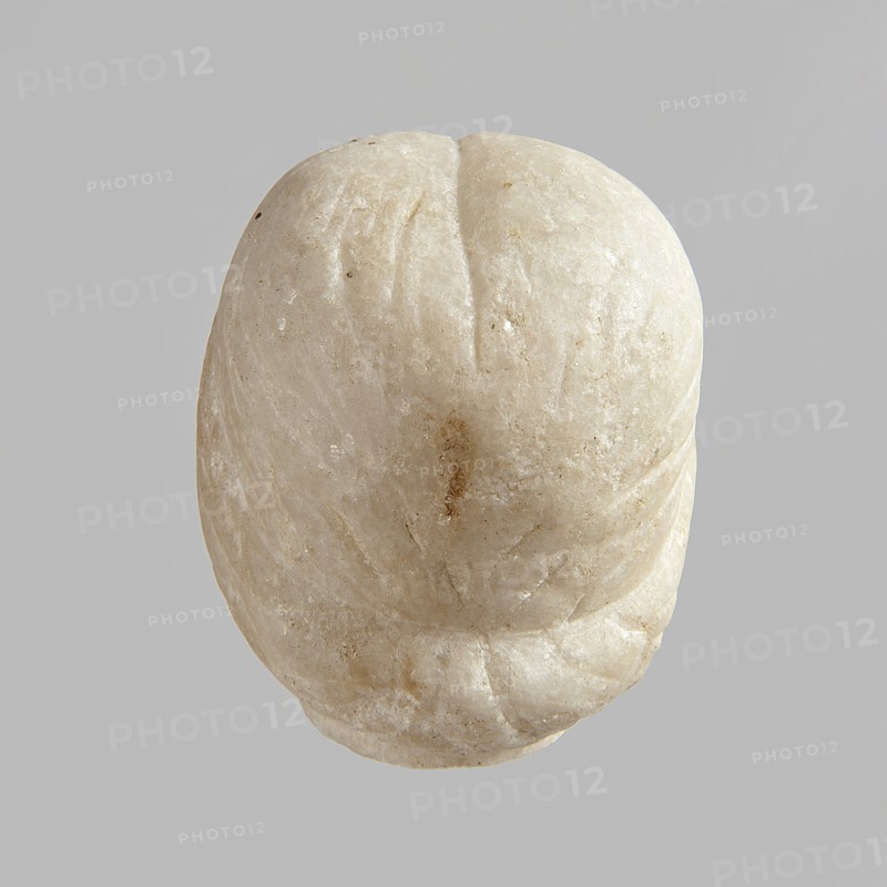 Fragment of a female figurine. Fragment of a small-sized sculpture or sculptural group with portrait features. The face is oval, the nose is large, the mouth is closed in a slight grimace. 2nd–3rd century AD, Egypt. Figurine, sculpture; height 8.5 cm, width 4.5 cm, depth 5.5 cm., A rounded stone object in off-white and beige tones sits against a flat gray background, showing a smooth, slightly translucent surface with small rough patches and areas of darker beige discoloration; the object has a central shallow groove and several faint curved indentations, with a lower edge that appears slightly chipped and irregular.