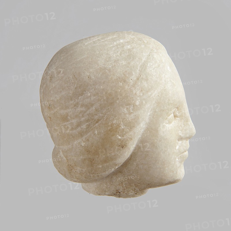 Fragment of a female figurine. Fragment of a small-sized sculpture or sculptural group with portrait features. Oval face, large nose, mouth closed in a slight grimace. 2nd–3rd century AD, Egypt. Terracotta figurine sculpture. Height 8.5 cm, width 4.5 cm, depth 5.5 cm., A carved stone head viewed in profile against a plain light gray background, showing a smooth, rounded top and stylized hair rendered with subtle grooves; the facial features include a pronounced forehead, a defined nose, gently closed lips, and a softly rounded chin. The surface of the stone displays a pale off-white to creamy beige coloration with variations of light tan and faint speckles, and areas of slight translucency and natural grain. The overall texture appears polished in places and slightly rough in others, with soft shadows accentuating the form.