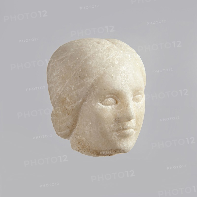 Fragment of a female figurine. Fragment of a small-sized sculpture or sculptural group. The depiction has portrait features. The face is oval, the nose is large, the mouth is closed in a slight grimace. 2nd–3rd century AD, Egypt. Figurine, sculpture; height 8.5 cm, width 4.5 cm, depth 5.5 cm., A pale stone sculpted head with smooth rounded contours, showing a stylized face with closed eye sockets, a defined nose and lips, and hair shaped close to the head; the stone is off-white to cream with subtle darker beige speckles and slight surface weathering, set against a uniform light gray background.