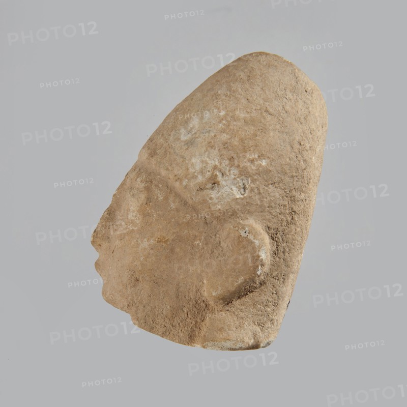 Head of a man, Archaic Period, Cyprus; figurine; height 7 cm, width 5 cm, depth 5.5 cm., A roughly triangular, weathered stone sculpture of a human head shown in profile against a plain light gray background, with a textured surface in shades of beige, tan, and light brown and areas of lighter whitish discoloration; the profile shows a pronounced forehead and sloping nose, an outlined ear, and a rounded chin, with natural pits and roughness visible across the form.