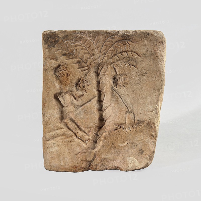 Hexagonal matrix for imprinting terracotta decorations, 1st–3rd century; Roman period, Tell Atrib, form; height 15.8 cm, width 15.3 cm, depth 12 cm, The image shows a rectangular carved stone tablet with a relief depicting a person reclining or leaning against a central palm-like tree with a textured trunk and radiating fronds; the person and tree are carved in shallow relief with visible tool marks and rough edges. The stone is mostly beige and light brown with some darker brown and grayish patches and worn areas; the background around the tablet is a plain light gray.