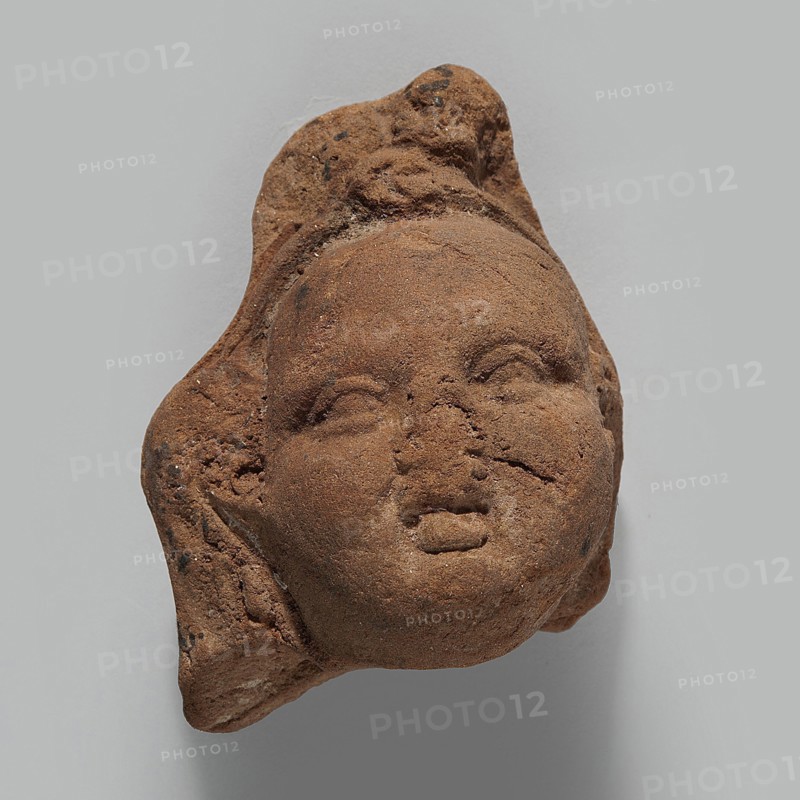 Head of Harpocrates; unknown creator; Harpocrates mythology; Hellenistic period, 332–30 BC, Egypt; figurine; height 5.4 cm, width 4 cm, depth 3.1 cm., A weathered clay or terracotta fragment shaped like a human head, with a rounded face, closed or partially closed eyes, a small nose, and slightly parted lips, showing rough, pitted texture and surface cracks; the piece has irregular edges and what appears to be a stylized hair or headdress form extending from the top and sides; colors visible include shades of brown, reddish-brown, and small darker brown or black flecks, all set against a neutral light gray background.
