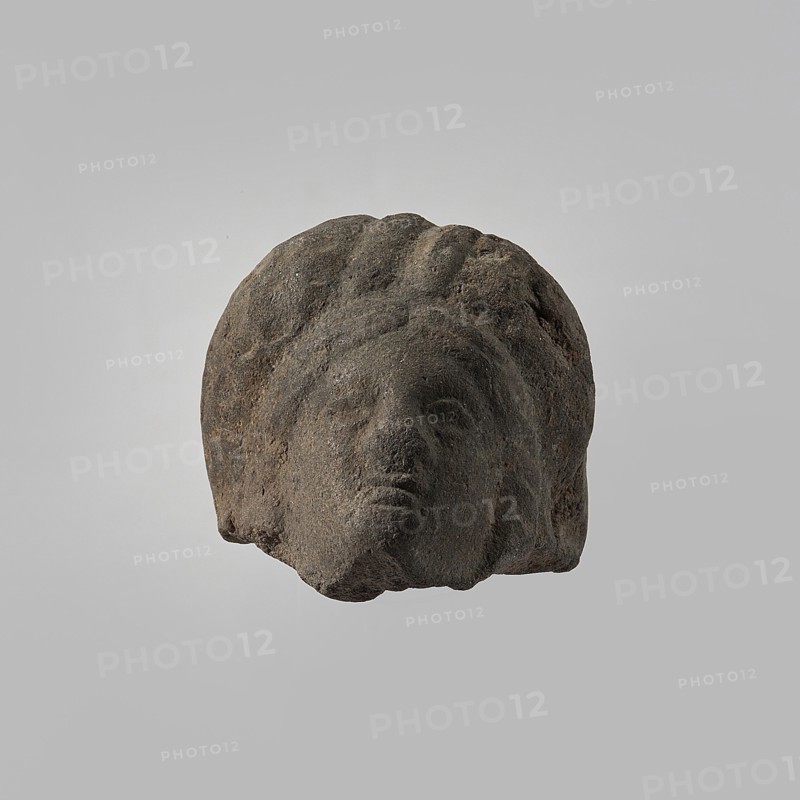 Head of a female figurine, preserved from the chin up. High hairstyle with a wreath above the forehead, composed of oblique, wide strands of hair. Schematic facial features, wide nose. Unworked back, smooth, rounded. Dark gray clay. 332–30 BC; Hellenistic period, Egypt. Figurine. Height 3.3 cm, width 3.7 cm, depth 3.5 cm., A weathered stone sculpture fragment showing a human head with stylized hair and facial features, rendered in dark gray and brown tones with a rough, pitted texture, set against a plain light gray background.