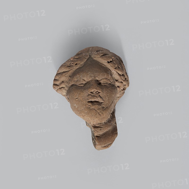 Head of a terracotta female figurine with part of the neck and a fragment of a robe preserved. Hairstyle parted in the middle. Facial features quite simplified and rounded. Light brown clay with uneven firing — center of the gray color break. Dated 332–30 BC, Hellenistic period, Egypt. Figurine; height 3.8 cm, width 3 cm, depth 2.1 cm., A weathered brown stone fragment shaped like a human face with stylized hair and softened facial features casting a faint shadow on a pale gray background, showing textured rough surfaces and areas of erosion with visible contours of a nose, closed mouth, and suggestion of eyes.