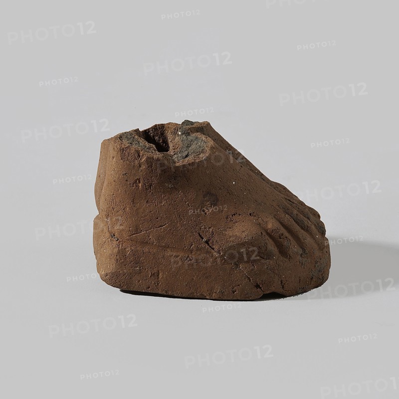 Fragment of a terracotta figurine showing two feet placed side by side on a flat, rounded base. Hellenistic or Roman period, Egypt, 323 BC–476. Figurine; height 4 cm, width 5 cm, depth 6.3 cm., A roughly foot-shaped object made of brown, earthy material sits on a light gray surface against a pale gray background; the object has a textured, uneven surface with a few small dark gray and black patches and some tiny lighter specks, visible depressions resembling toes and a hollowed top, and casts a soft shadow beneath and slightly to the right.