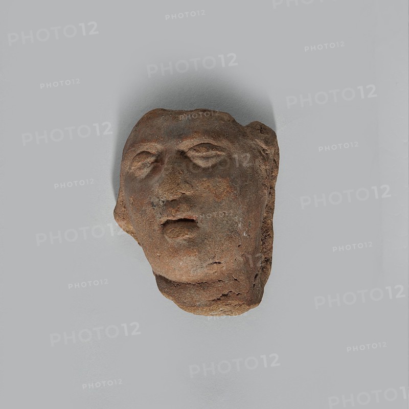 Fragment of the head of a terracotta female figurine. Fragmentarily preserved head depicting a woman: face and upper part of the neck. Narrow, open mouth with wide lips; large eyelids giving the eyes a half-closed appearance; fairly wide nose. Hairstyle unpreserved. Made of fairly poor quality clay with minor cracks especially on the neck and chin and visible grains; surface slightly effaced. Hellenistic or Roman period, Egypt, 323 BC–476 AD. Figurine; height 3.6 cm, width 3.0 cm, depth 2.3 cm., A worn sculpted head fragment in shades of brown and reddish-brown rests against a light gray background; the fragment shows an abstracted human face with closed or partially closed eyes, a nose, and a slightly open mouth, and the surface displays rough, pitted texture with areas of darker and lighter brown.