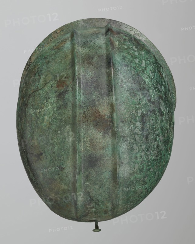 Bronze helmet with rectilinear cutouts for the eyes, nose, mouth, and chin; the helmet has two prominent reinforcements ribs. 550–500 BC; Italy . Armament. Height 26 cm, width 19.3 cm, depth 25 cm, weight 0.994 kg., An oval, helmet-like object with a textured, mottled surface showing shades of green, turquoise, brown, and gray, featuring three raised ridges running lengthwise, a rounded rim with a row of small circular impressions along its edge, and a short metal peg protruding from the bottom; the background is a smooth pale gray.