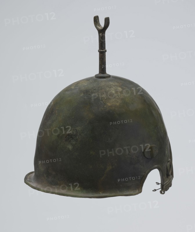 Bronze helmet, 4th–3rd century BC, Italy, armament; height 27 cm without cheek guards, width 19.8 cm, depth 24 cm, weight 0.738 kg without cheek guards., A rounded metal helmet with a smooth domed surface showing a patina of dark green, olive, brown, and muted gold tones, topped by a vertical metal rod ending in a forked shape, with a flared rim at the front and a cutaway section at the base on one side that includes a small metal clasp and looped fasteners; the background is a pale, light gray.