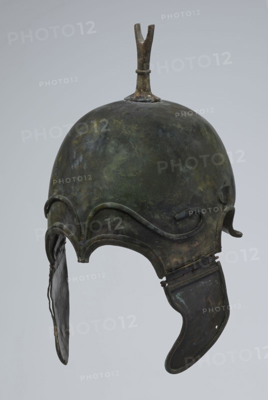 Bronze helmet, 4th–3rd century BC, Italy, armament; height 34 cm, width 20.2 cm, depth 22.5 cm, weight 1.035 kg., A metal helmet with a rounded dome and a small forked projection on top, showing a mottled surface of green, dark green, brown, and black patina; decorative raised curving ridges around the brow and cheek areas; a hinged cheek guard on one side with a curved lower edge and a hanging flap on the other side with a thin strip connecting it; overall worn and aged texture with uneven coloration against a pale gray background.