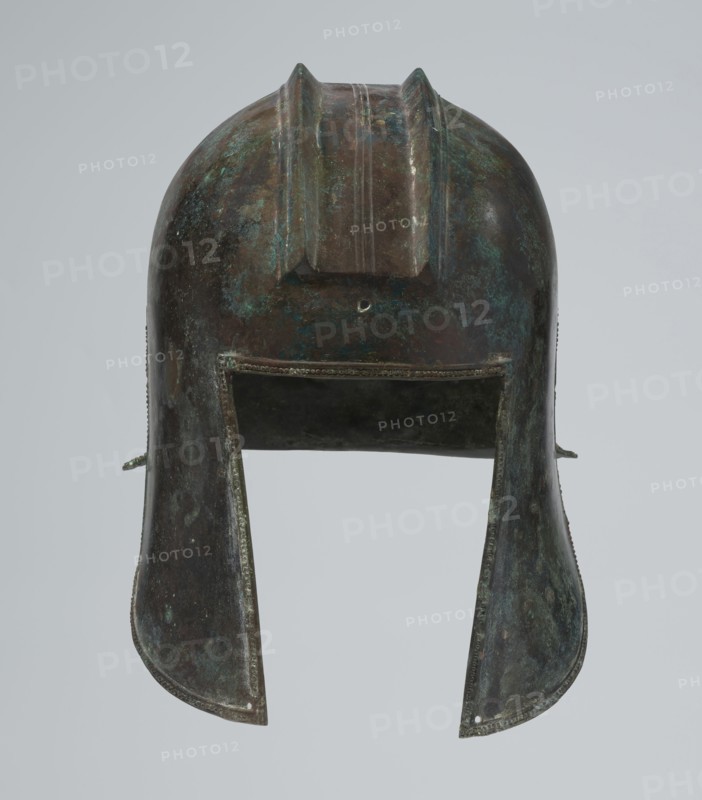 Bronze helmet, circa 550–500 BC, possibly Italy; armament. Dimensions: height 24 cm, width 18.5 cm, depth 23.2 cm; weight 1.170 kg., A single metal helmet viewed from the front with a rounded top and a raised central crest, showing a patina of greenish-blue and dark brown on its surface; a rectangular front opening leads into a darker interior, and two flared cheek guards extend downward on either side, each edged with a beaded trim and showing areas of mottled green-blue and brown; the background is a plain pale gray.