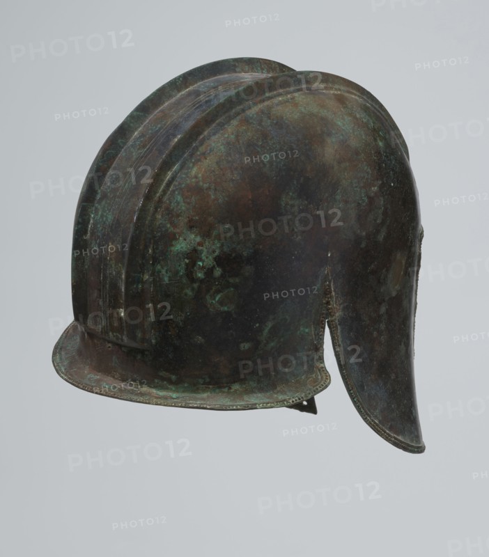 Bronze helmet, 550–500 BC, Italy, armament, height 24 cm, width 18.5 cm, depth 23.2 cm, weight 1.170 kg, A metal helmet with a rounded dome and a rear neck guard, showing a mottled surface of green, brown, dark bluish, and black tones, with areas of lighter green patina and darker brown spots, and a curved brim along the front edge.
